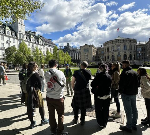 The group is gathered on a paved area, looking towards the Stortinget (Norwegian Parliament) building, which has a distinct rounded facade and a flag flying on its roof. On the left, a grand white building with a classic architectural style is visible. The scene is outdoors on a bright day with a partly cloudy sky.