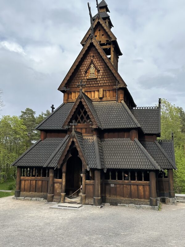 The intricately designed Gol Stave Church, a historic wooden church with tiered roofs and detailed carvings, stands outdoors under a cloudy sky. The dark wood of the church contrasts with the green trees visible in the background.