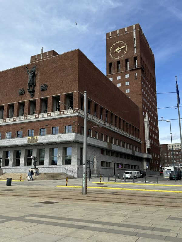 The distinctive brick facade of Oslo City Hall (Oslo Rådhus) is visible under a clear sky. The building features two large towers, with a clock face prominently displayed on the taller, right-hand tower. Tram tracks and a street are in the foreground, and a few people can be seen near the building's entrance.