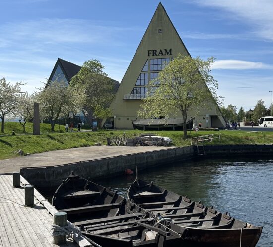 The pyramid-shaped Fram Museum building, with "FRAM" written on its facade, stands prominently on a sunny day. In the foreground, two traditional wooden boats are docked at a pier, with calm water next to them. Trees with green leaves and a clear sky are visible in the background, along with a road where a bus can be seen.