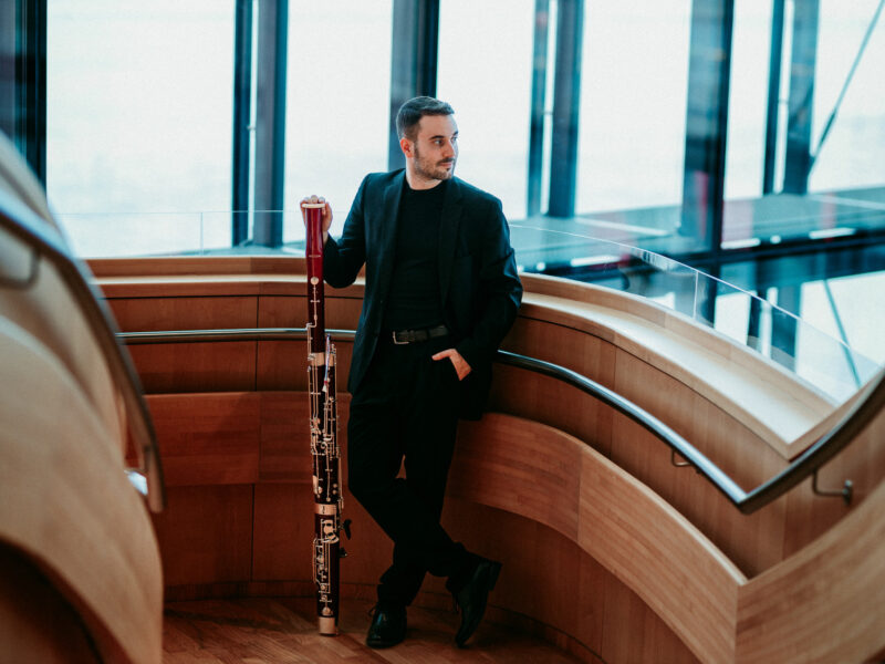 Alexandre Molina, dressed in a black suit, stands leaning against a light wooden railing, holding a bassoon. He looks to the side and light streams in through the large glass windows visible in the background.