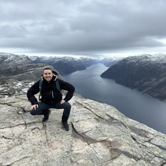 A young Institut Nòrdic student named Alexandre Molina smiles at the camera as he poses squatting in Preikestolen. He wears a black jacket and dark pants. Behind him stretches a breathtaking fjord and snow-capped mountains under the cloudy Norwegian sky.