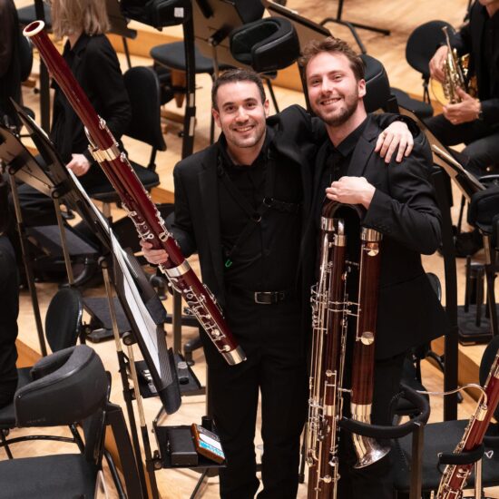 Alexandre Molina, a student at Institut Nòrdic, is pictured with another musician on stage at the Stavanger Symphony Orchestra. Both are wearing black suits, holding bassoons and smiling at the camera.