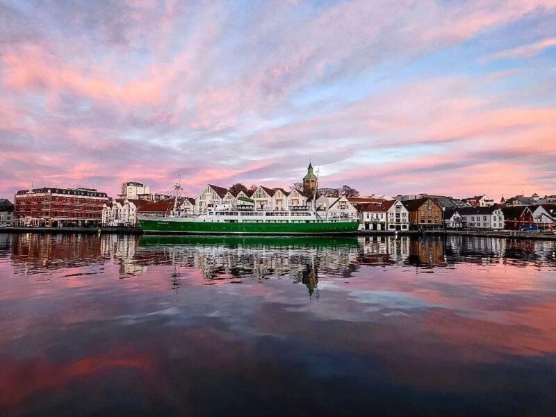 A view of Stavanger at sunset, where a large green ship floats in the calm waters of a harbor. Around the harbor are traditional red and white-roofed buildings, and the sky is tinged with shades of pink and blue.