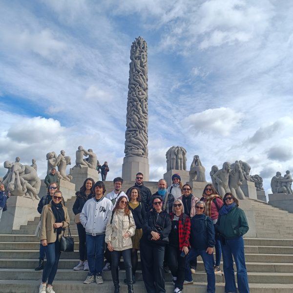A group of 17 students and two instructors from Institut Nòrdic are standing on stone steps in front of the Monolith, a tall, intricately carved sculpture by Gustav Vigeland, located within Vigeland Sculpture Park in Oslo. Other large stone sculptures are visible on either side, under a partly cloudy sky.